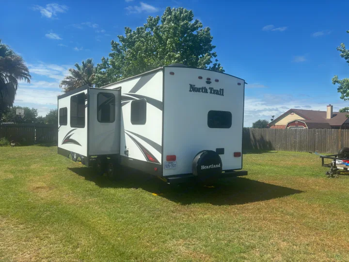 A white North Trail travel trailer, ideal for an RV rental, parked in a sunny Texas backyard with palm trees and a fence.. Heartland RVs North Trail 2018