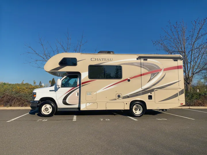 Beige Chateau RV rental parked in a lot under clear blue sky. This motorhome has an awning & ladder visible from the side view.. Thor Motor Coach Chateau 2017
