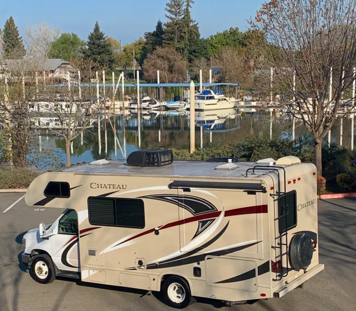 Tan Class C RV rental parked near a calm marina with various boats docked, offering a scenic view.. Thor Motor Coach Chateau 2017