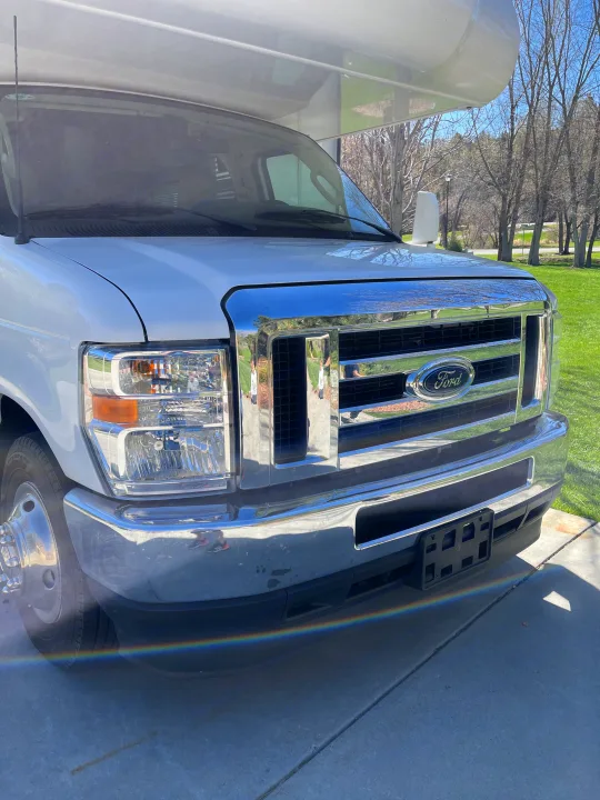 Front of a white Ford RV rental. Sunlight glints off the chrome grille, with a rainbow visible on the bumper.. Thor Coleman 2024