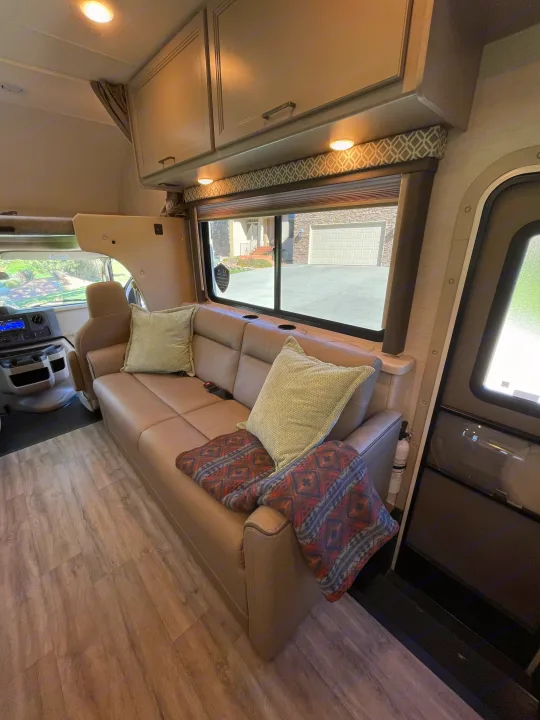 Inviting RV rental interior: beige sofa with pillows, wood floor, overhead cabinets, and large window overlooking a driveway.. Thor Coleman 2024