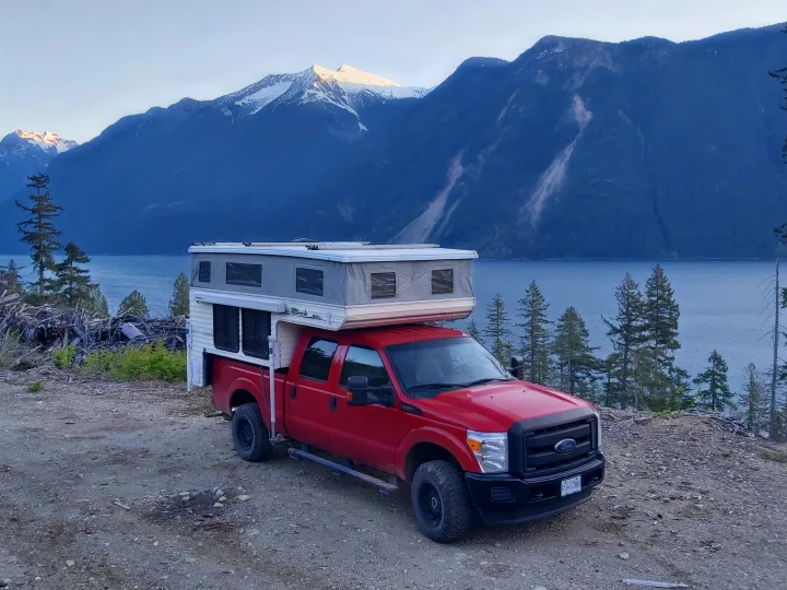 Red pickup truck with a pop-top camper, perfect for an RV rental, overlooking a scenic mountain lake with snow-capped peaks.. Ford F350 2015