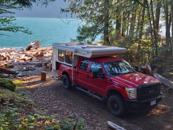 Scenic rv rental: Red Ford truck with a Hallmark pop-top camper parked by a driftwood-laden beach with blue water.. Ford F350 2015