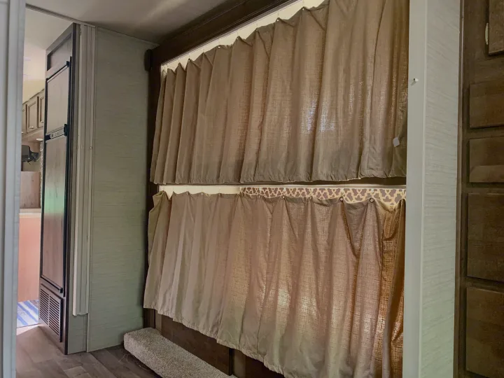 Inside an RV rental: two levels of light privacy curtains for bunk beds, with a refrigerator and cabinets visible on the sides.. Jayco Redhawk 2019