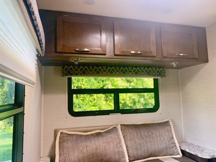 Cozy RV rental bedroom features overhead cabinets, a window with lush green views, textured walls, and comfy pillows.. Jayco Redhawk 2019
