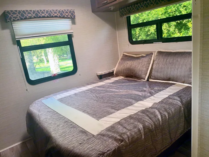 Experience comfort in this RV rental bedroom featuring a full bed with grey bedding, dual windows for scenic views, and ample natural light.. Jayco Redhawk 2019