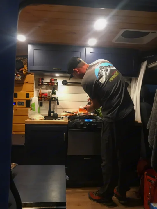 Man cooks in a well-equipped RV rental kitchen. Gas stove, sink, and storage make mobile living easy for travel.. ram promaster 2020