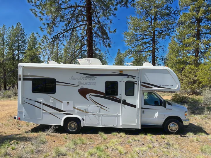 A white Jamboree Searcher RV rental is parked among tall pine trees on a sunny day, perfect for a nature getaway.. Jamboree Searcher 2013