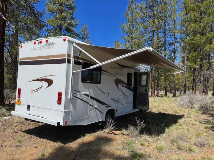 Spacious Jamboree Searcher RV rental with extended awning, parked in a sunlit pine forest. Perfect for camping.. Jamboree Searcher 2013