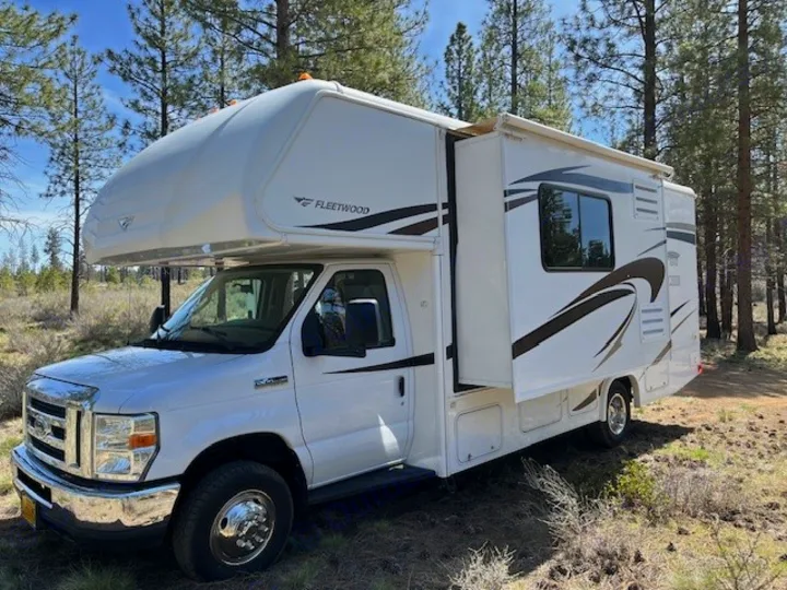 Explore nature with this clean white Fleetwood RV rental, parked in a sunny forest setting with its slide-out extended.. Jamboree Searcher 2013