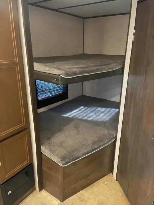 An RV rental's sleeping area, featuring cozy bunk beds with grey mattresses, a window, and nearby cabinetry for convenience.. Coleman 262BH 2021