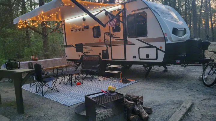 A well-appointed RV rental travel trailer campsite at dusk, with glowing awning lights, an outdoor rug, and a fire pit.. Winnebago Minnie Winnie 2021