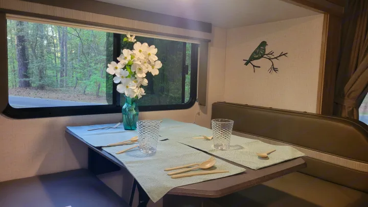 RV rental dinette with a forest view, table set for two with placemats, glasses, and a vase of white flowers.. Winnebago Minnie Winnie 2021