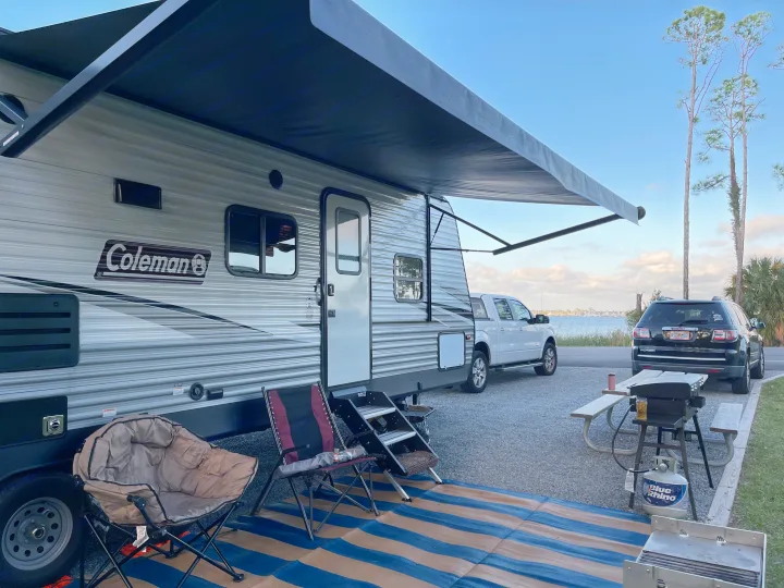 A Coleman RV rental is set up at a scenic waterfront campsite with an extended awning, chairs, and a grill.. Coleman 262BH 2021