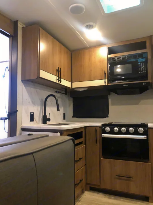 Modern rv rental kitchen with light wood cabinets, black appliances (microwave, stove), and a sink, offering a compact cooking area.. Grand Design Imagine 2023