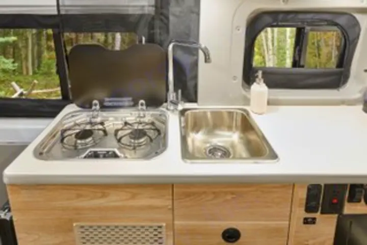 This RV rental features a compact kitchen with a two-burner stove, sink, and light wood cabinetry. Forest scenery visible through windows.. Winnebago Solis PX 2025
