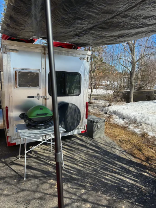 RV rental setup: A silver trailer with a green grill, cooler, and tarp awning. Parked in a snowy, wooded area for outdoor adventure.. Weeroll Silver cloud 2020