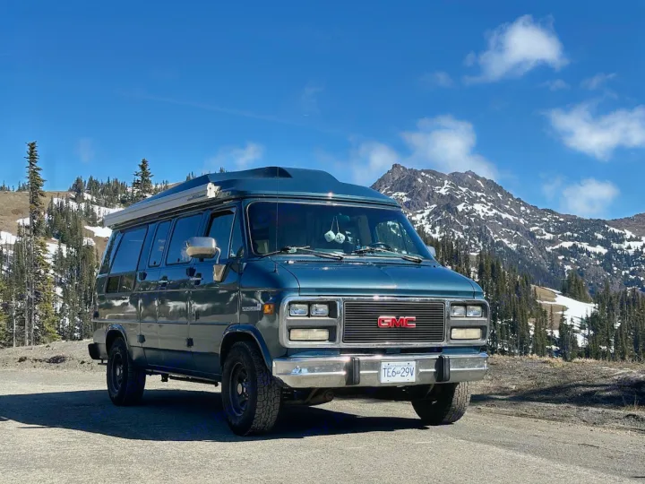 A teal GMC camper van, ideal for an rv rental, parked on a scenic mountain road amidst snow-dusted peaks.. GMC Vandura 2500 1992