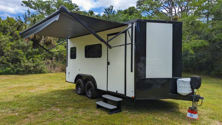 Modern white & black toy hauler/travel trailer with extended awning. An ideal RV rental unit, parked on grass by green trees.. Diamond Cargo 16x8.5 2023