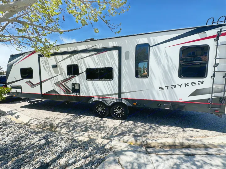 White Stryker toy hauler RV travel trailer parked outdoors on a sunny day with tree shadows. Consider for an RV rental!. Cruiser Rv Corp Stryker 2021