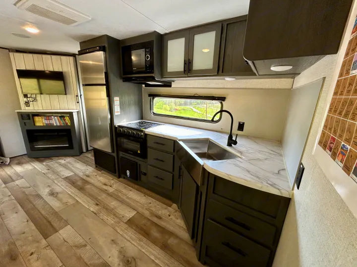 Modern RV rental kitchen & living space. Features marble counters, black appliances (sink, stove, microwave, fridge), TV, and fireplace.. Aspen Trail 3280BHS 2022