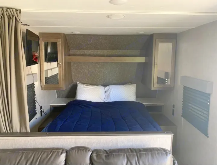 Inside an RV rental: a compact bedroom with a bed, navy comforter, wall-mounted TV, and integrated storage.. Shasta Oasis 2021
