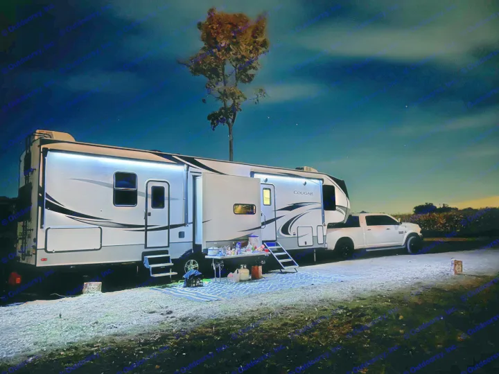 Cozy night at an RV rental campsite. Brightly lit fifth-wheel and truck with outdoor setup, perfect for relaxation.. Keystone RV Cougar 2021