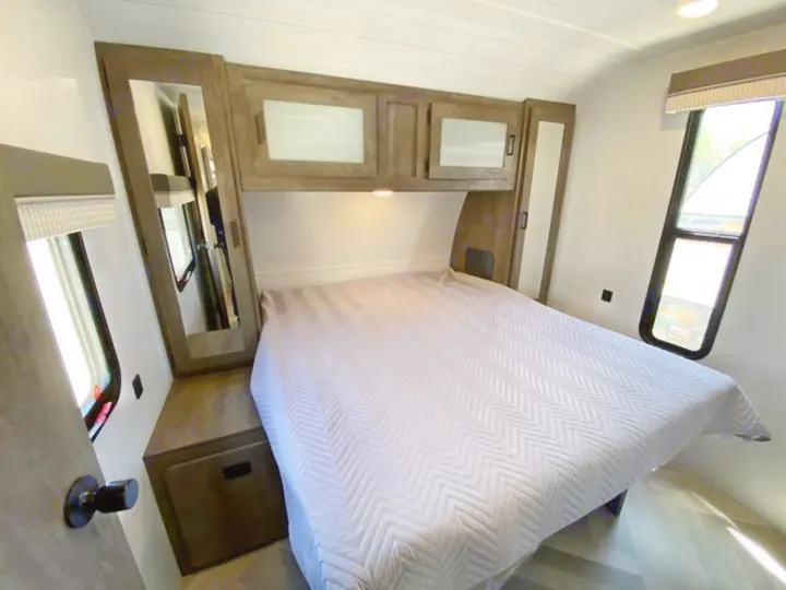 RV rental bedroom: a comfortable space featuring a queen bed, overhead wood cabinets, and large windows.. Forest River Wildwood 2021