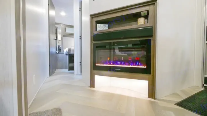 Cozy RV rental interior with modern electric fireplace & built-in shelving. Hallway leads to the bedroom with herringbone floors.. Forest River Wildwood 2021
