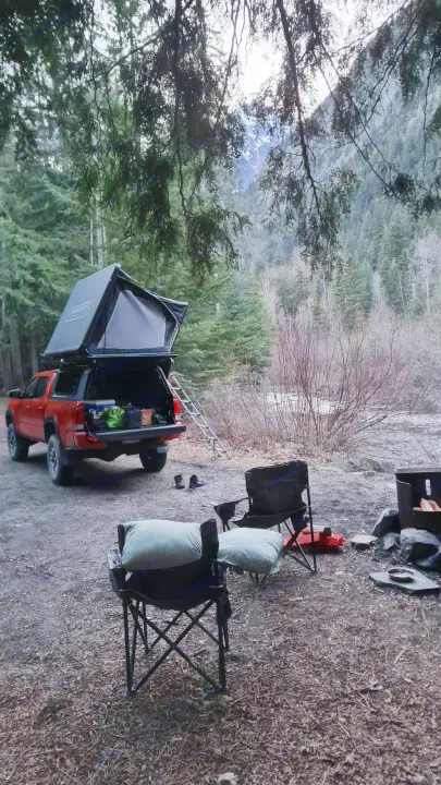 Forest campsite: orange truck with rooftop tent, chairs, and fire pit by a river. An RV rental offers similar outdoor adventures.. Toyota Tacoma TRD OFF ROAD 2018