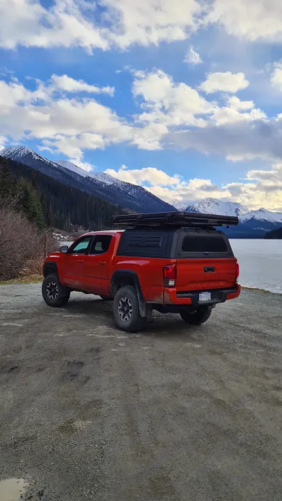 A red pickup truck with a rooftop tent parked by a frozen lake, surrounded by snowy mountains and pine forests. Perfect for an RV rental adventure.. Toyota Tacoma TRD OFF ROAD 2018