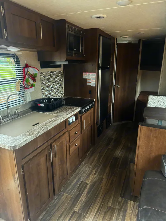 Inside an rv rental: a kitchen with dark cabinets, stainless sink, microwave, fridge, stove, and a dining table with wood-look floors.. Shasta 26db 2020