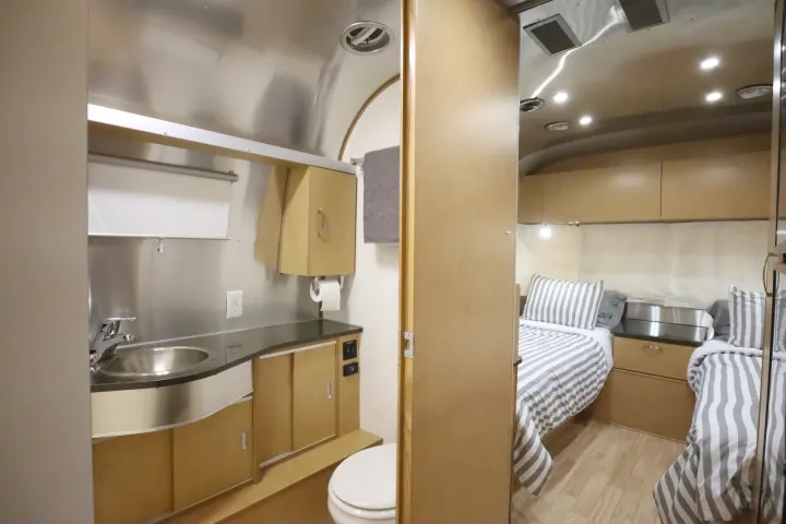 This modern RV rental interior features a sleek bathroom with stainless sink & toilet, next to a bedroom with striped bedding.. Airstream Flying Cloud 2016
