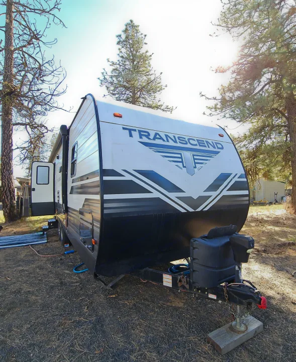 An RV rental: a white and black Transcend travel trailer parked at a sunny gravel campsite, surrounded by tall trees.. Grand Design Transcend 2019