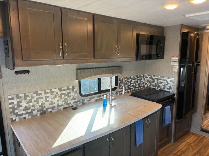 Modern RV rental kitchen with dark cabinets, light counters, mosaic backsplash, stainless sink, microwave, and fridge.. Nexus RV Triumph 2021