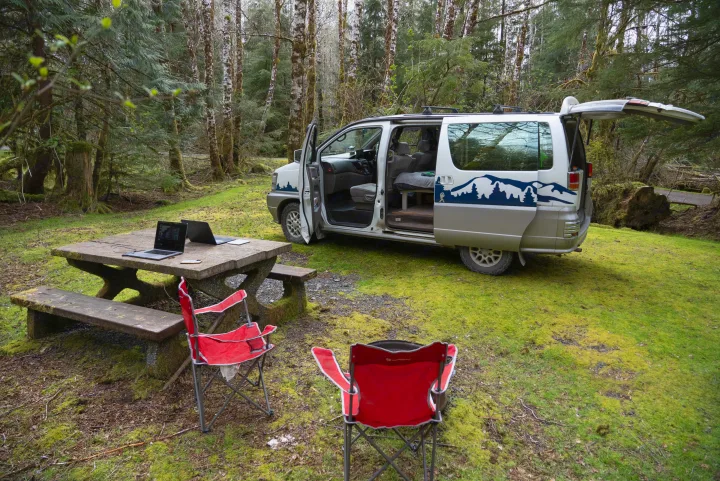 An RV rental campervan with open doors sits in a green forest. A laptop on a picnic table & red chairs form an outdoor office.. nissan elgrand 1997