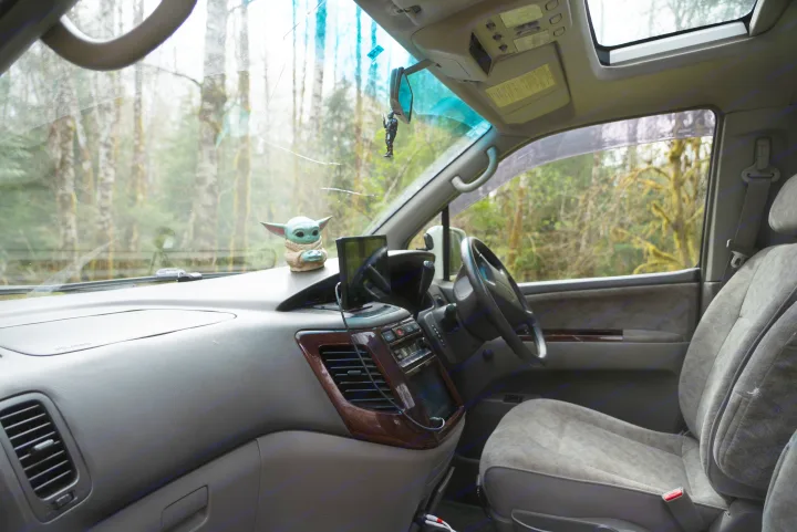 Inside an RV rental: dashboard with Grogu figurine, Mandalorian charm, and a view of trees through a cracked windshield.. nissan elgrand 1997