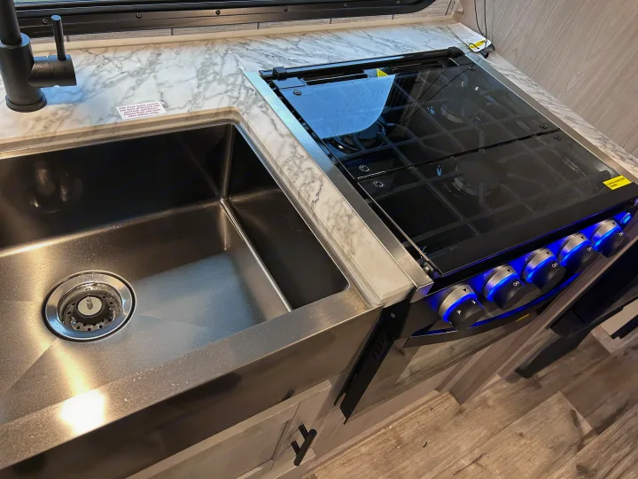 Modern RV rental kitchen featuring a sleek stainless steel sink, dark faucet, and a black glass stovetop with blue-lit controls.. 4YDTCMH25RJ935536 1805RB 2024