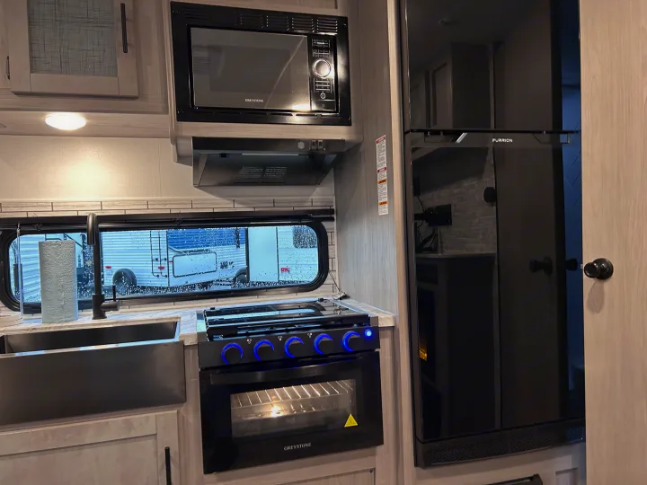 Inside a modern RV rental kitchen featuring a Greystone microwave/oven, Furrion fridge, sink, and lit stovetop.. 4YDTCMH25RJ935536 1805RB 2024