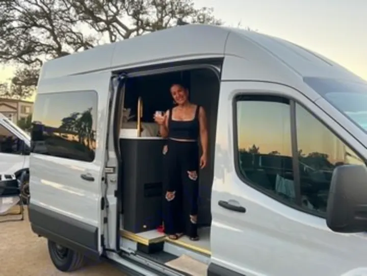 Smiling woman inside a sleek gray **RV rental** **campervan**, holding a drink, ready for **travel** and **adventure**.. Ford Transit 2023