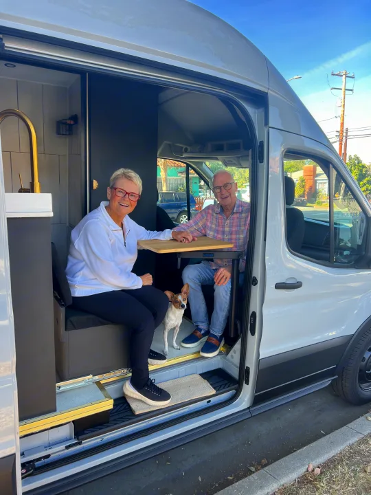 Inside a bright RV rental van, a smiling couple and their dog relax at a table, ready for travel.. Ford Transit 2023