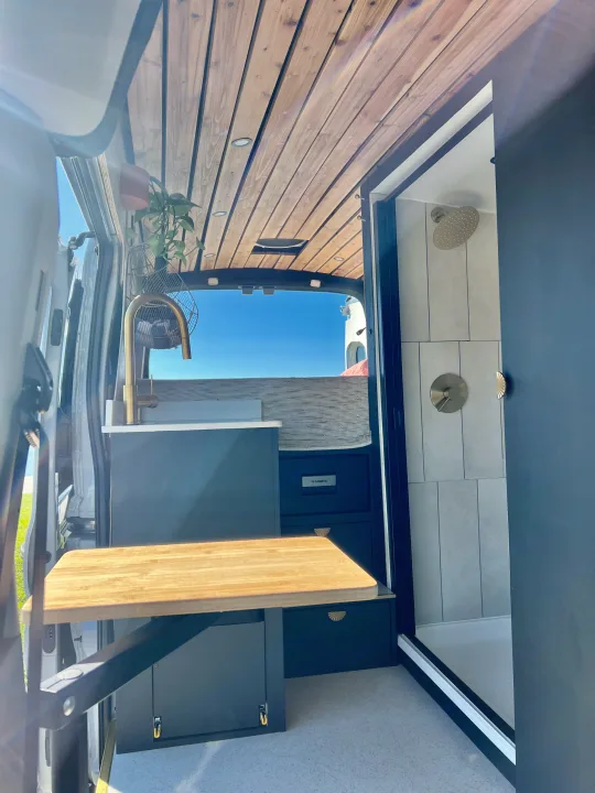 Modern rv rental interior: stylish kitchenette with golden faucet, wood ceiling, fold-out table, and full shower with elegant fixtures.. Ford Transit 2023
