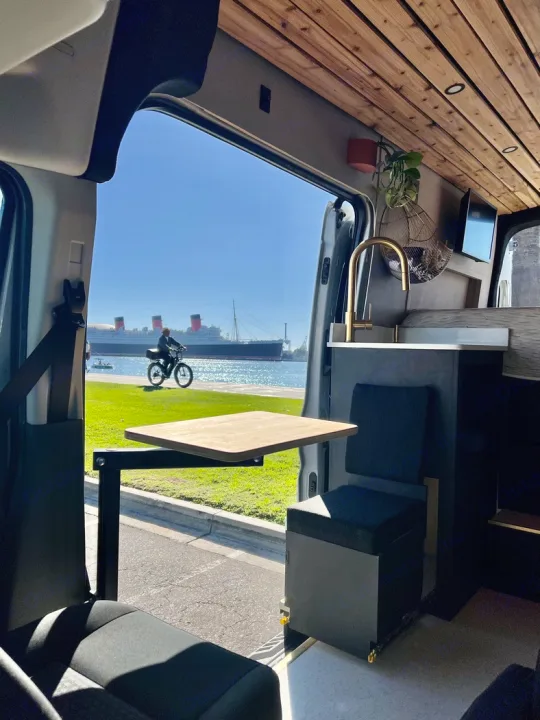 RV rental interior with wooden ceiling, table, and sink. Enjoy scenic views of the Queen Mary and bay from this camper van.. Ford Transit 2023