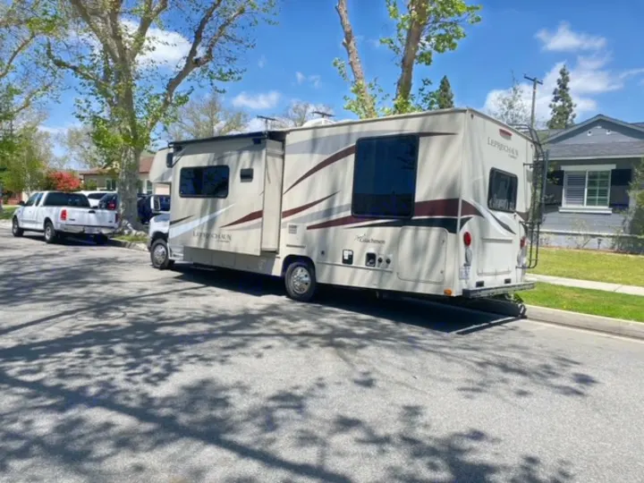 A beige Coachmen Leprechaun RV rental is parked on a sunny residential street, with a white pickup truck nearby.. Coachmen Leprechaun 2017