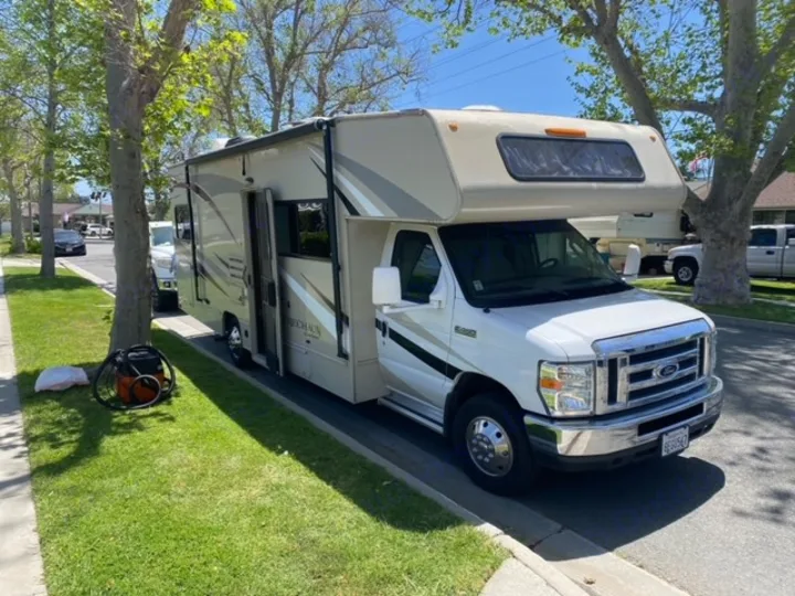 White and beige Class C **RV rental** parked on a sunny residential street. A vacuum sits nearby, suggesting prep work.. Coachmen Leprechaun 2017