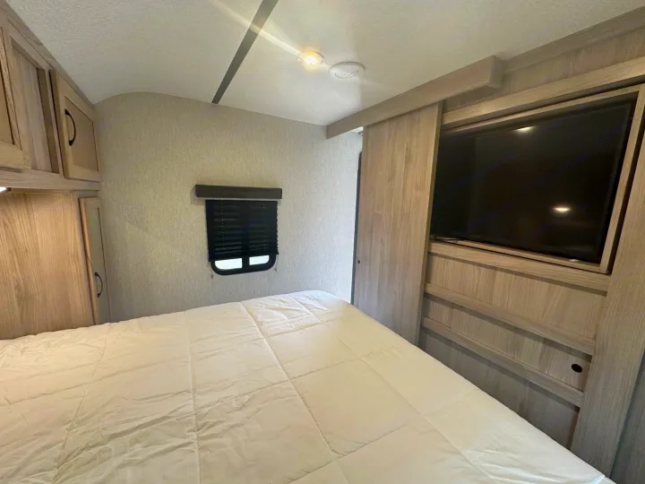 This RV rental bedroom features a white bed, wall-mounted TV, and light wood cabinetry, creating a compact and functional space.. coachman spirit 2022
