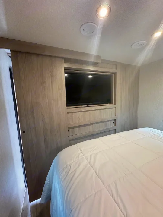 RV rental bedroom with a white comforter on the bed, a wall-mounted TV in built-in wood cabinetry, and a sliding door.. coachman spirit 2022