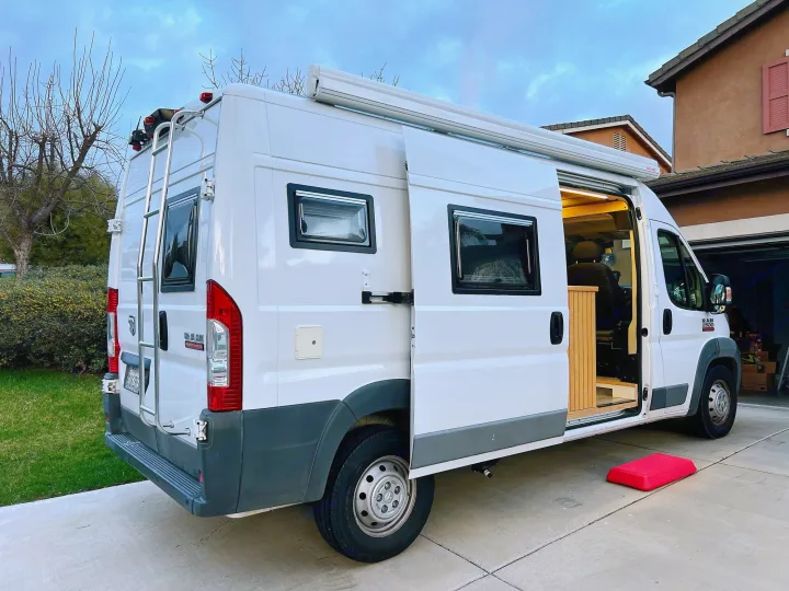 White camper van **rv rental** with open side door, interior lights, & awning extended. Rear ladder visible.. Ram promaster 2500 2017