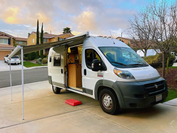 A white camper van RV rental, with its awning extended, sits on a driveway revealing a custom wood interior.. Ram promaster 2500 2017