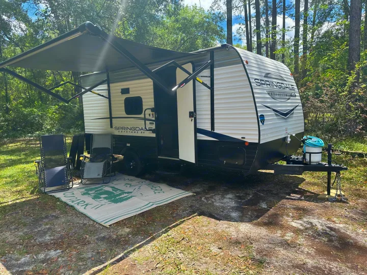 Getaway with an RV rental! White travel trailer with extended awning, outdoor rug & chairs at a serene wooded campsite.. Keystone RV Springdale 2024